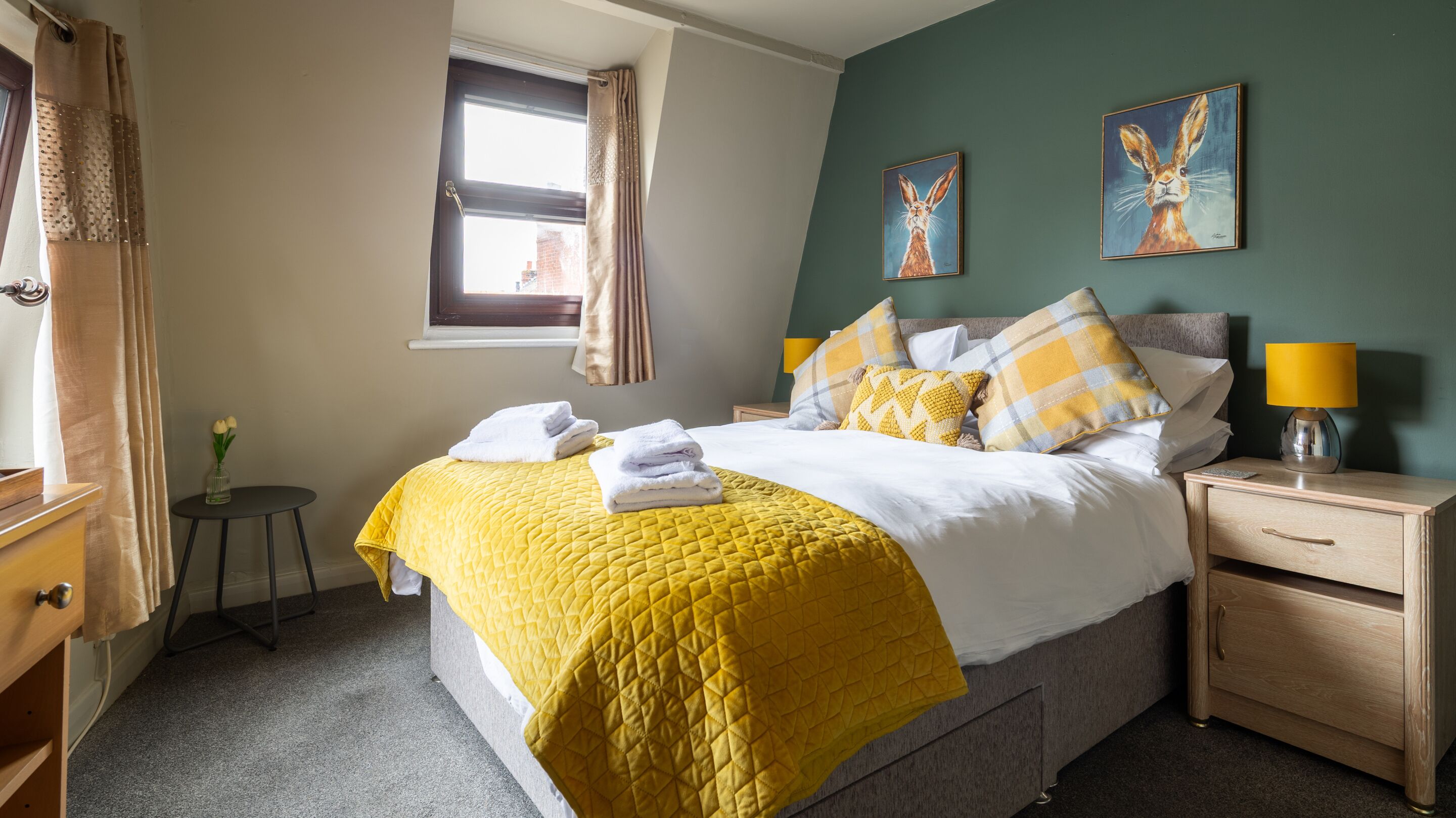 A neatly made bed with a yellow quilt, a bedside lamp, and two paintings on the wall.