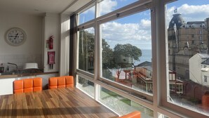 A modern kitchen with a wooden table and orange chairs, large windows with a view of buildings and trees, and a wall clock. - Harcourt Place (Scarborough)
