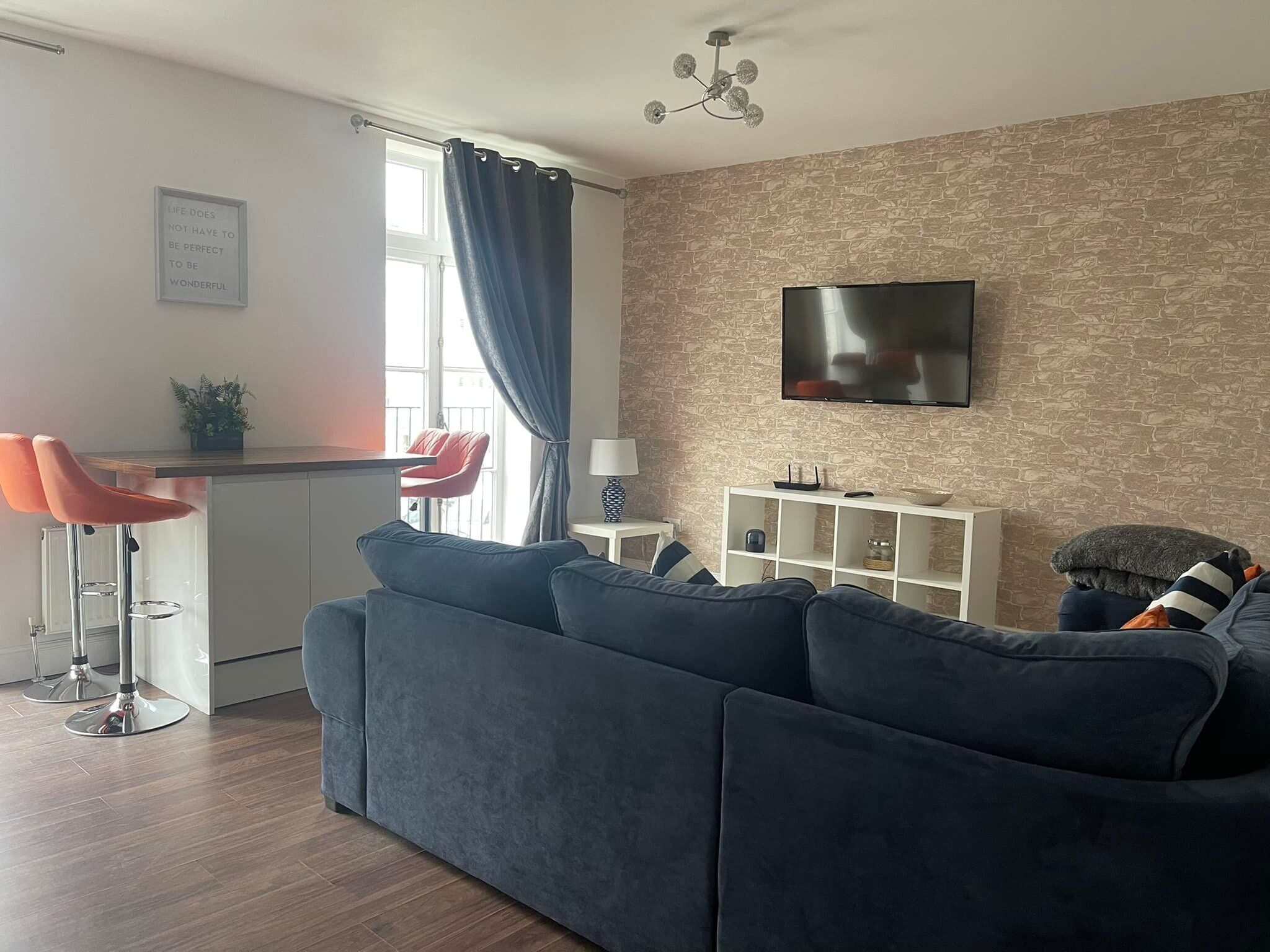 Una sala de estar moderna con un sofá azul oscuro, una silla roja, una zona de cocina con barra y una pared con textura de piedra.