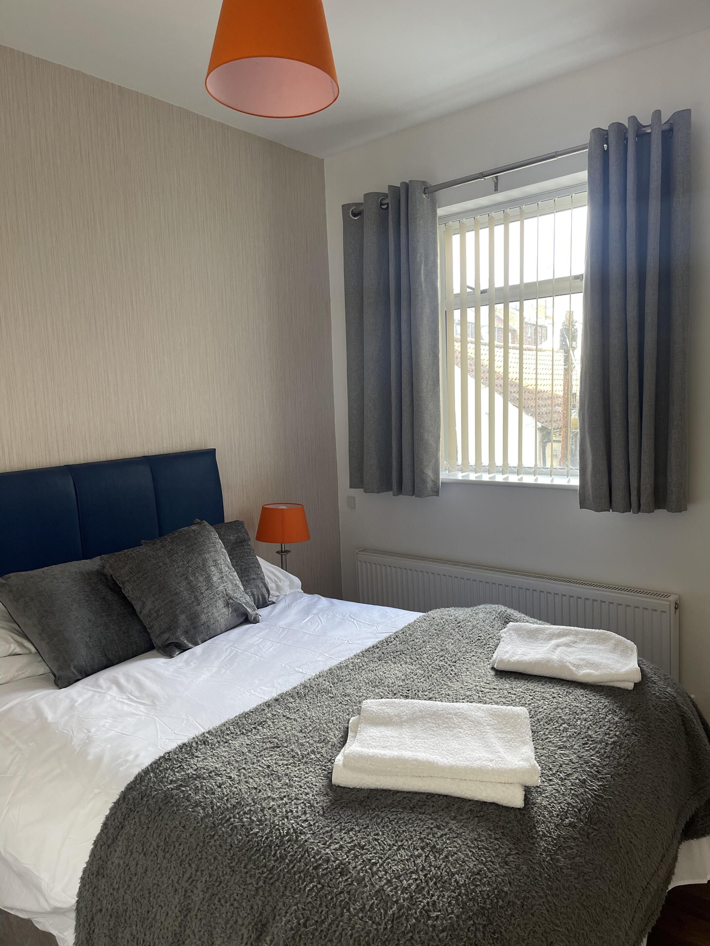 A neatly made bed with a grey blanket and two white towels, a blue headboard, a window with grey curtains, and a lamp on the nightstand.