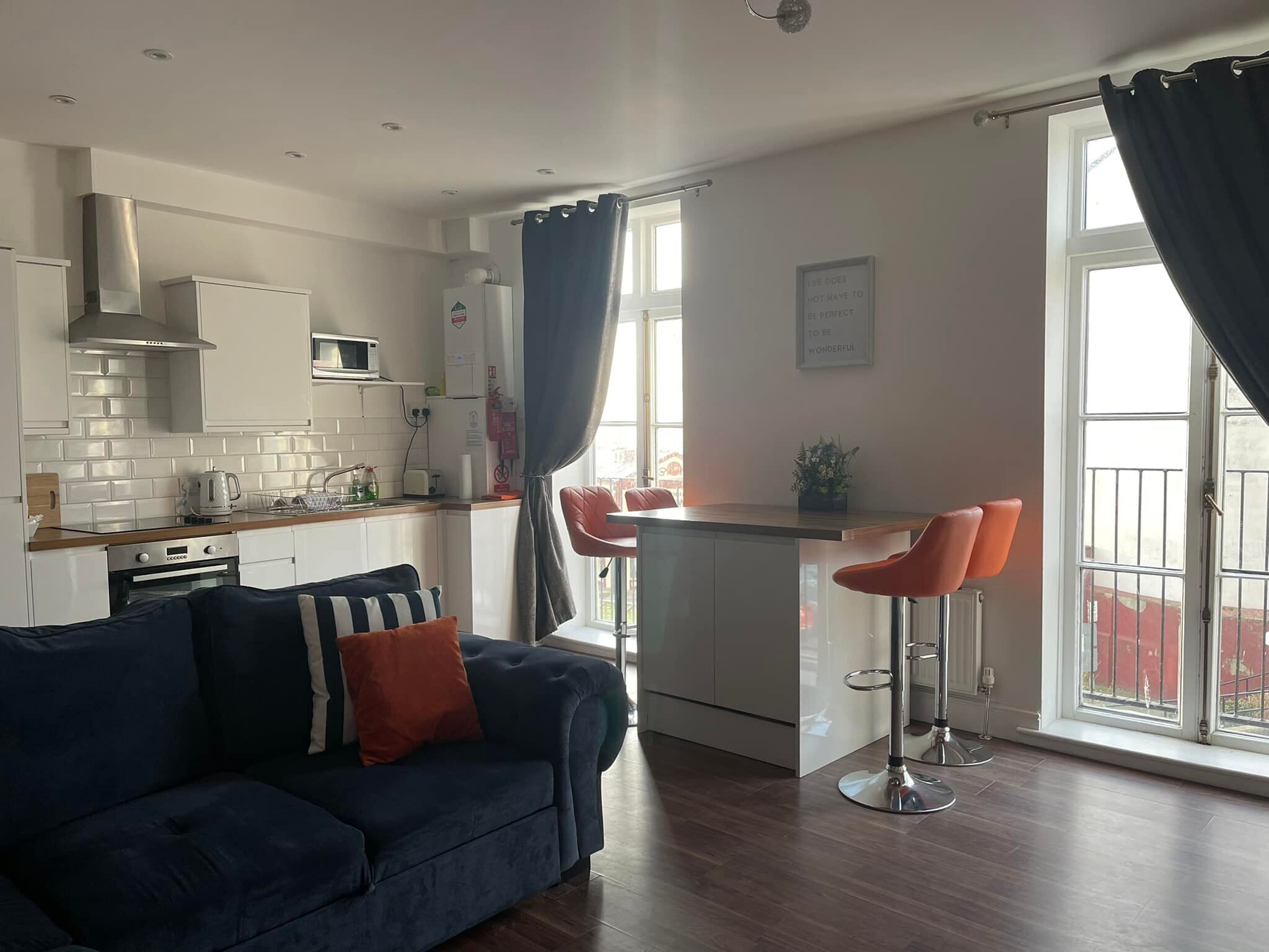 A compact living space with a kitchenette, a sofa, a dining area with two bar stools, and large windows.