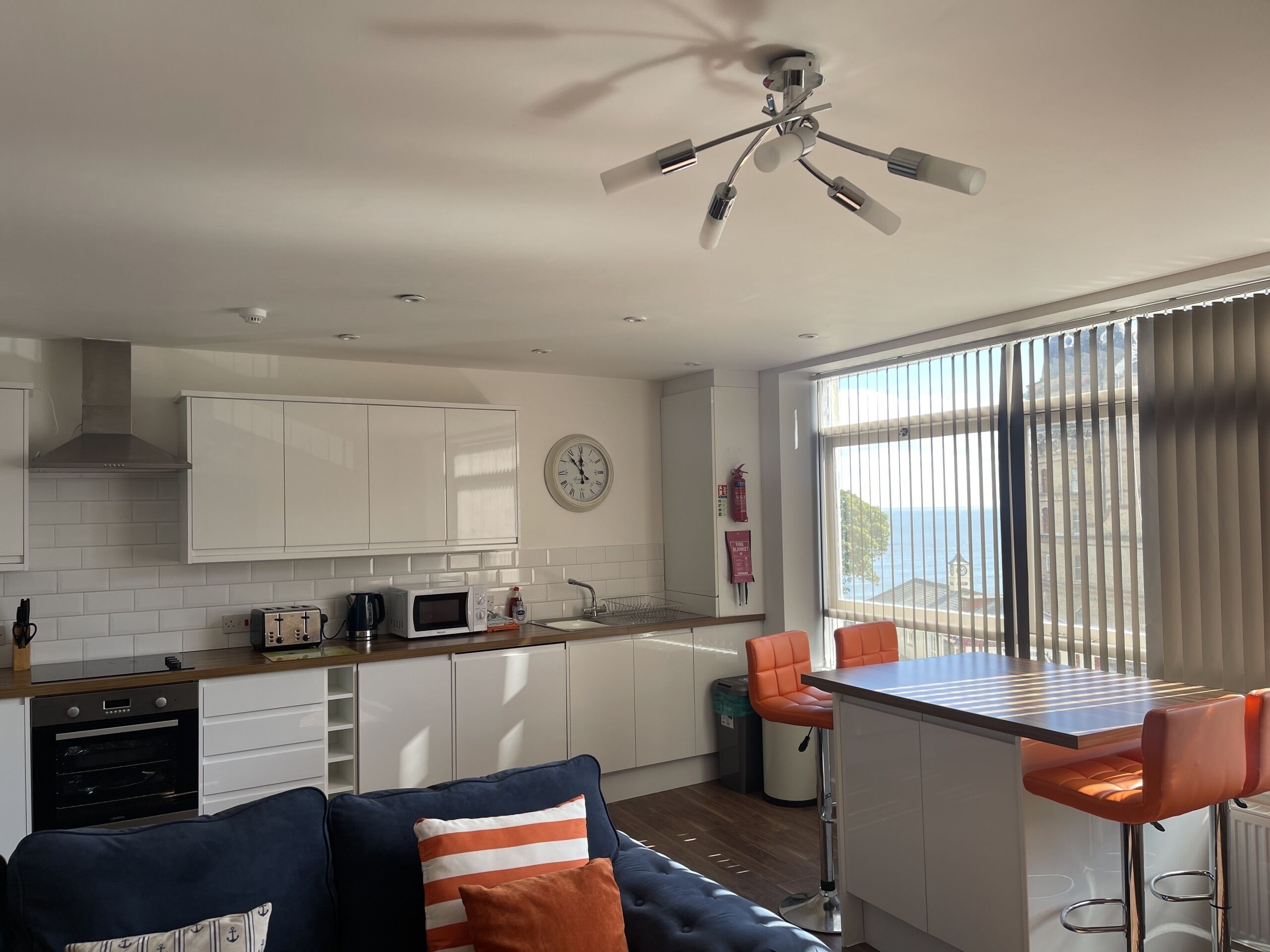 Cocina moderna con armarios blancos, microondas y cafetera. Un comedor con una mesa y sillas naranjas. Un sofá azul con almohadones a rayas se encuentra en primer plano.