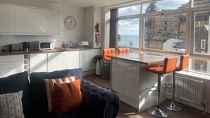A modern kitchen with white cabinets, a microwave, and a toaster. There is a large window with a view of the sea and buildings. - Harcourt Place (Scarborough)