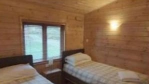 A wooden cabin room with two beds, a window, and a wall-mounted light. - The Log Cabin Glebe Farm (Market Rasen)