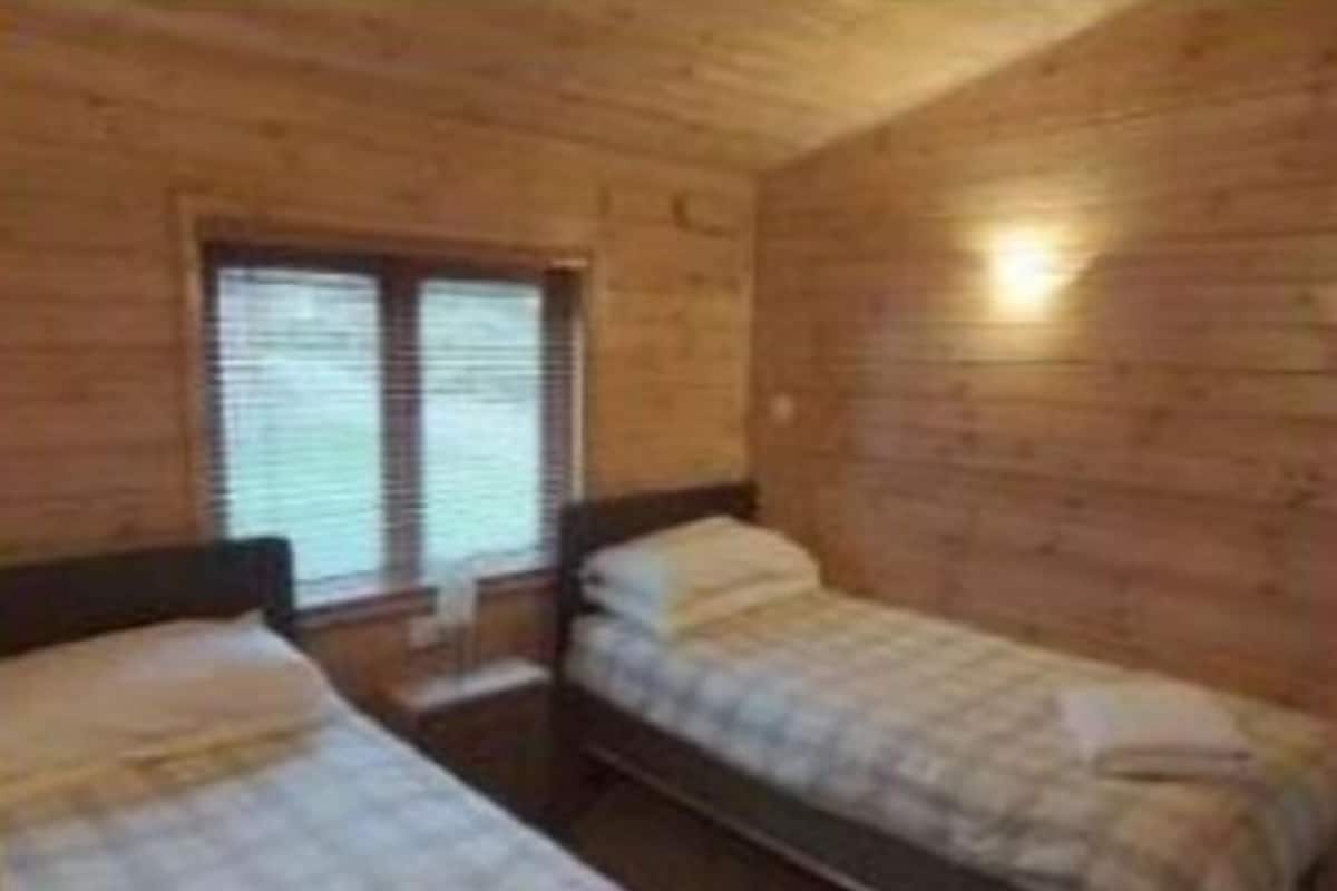 A wooden cabin room with two beds, a window, and a wall-mounted light.