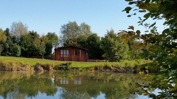A cabin by a lake with a reflection, surrounded by trees and grass. - The Log Cabin Glebe Farm (Market Rasen)