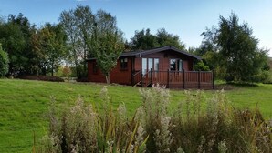 A wooden cabin with a deck, surrounded by a grassy field and trees. - The Log Cabin Glebe Farm (Market Rasen)