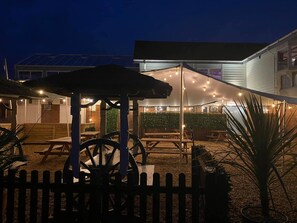 An outdoor patio area with wooden tables and chairs, a covered seating area, and string lights.