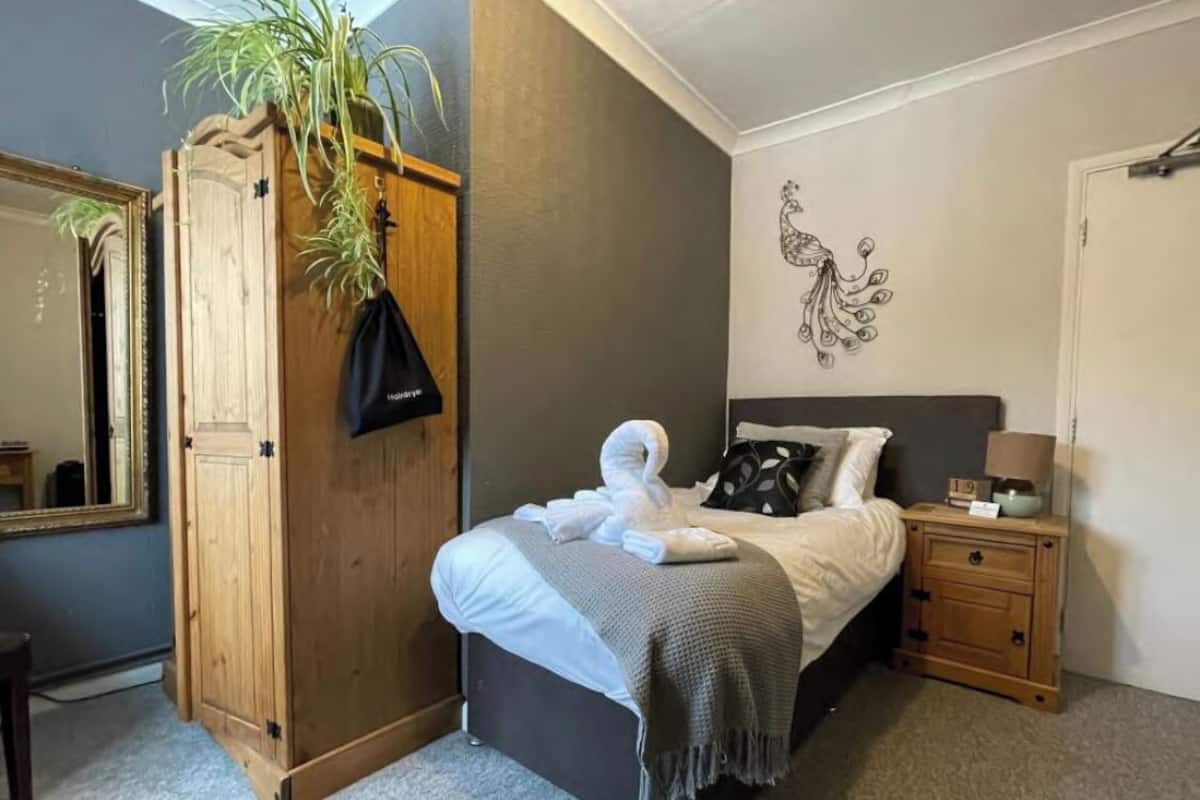 A bedroom with a bed, a wooden wardrobe, a mirror, a wall decoration, and a nightstand.