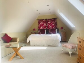 A bedroom with a sloped ceiling, a bed with white bedding, a red floral curtain, a glass coffee table, a wicker chair, and a wooden dresser. - Rosebank Bed & Breakfast (Etchingham)
