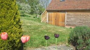 A wooden barn with a sloped roof and a wooden door, surrounded by a grassy area with flowering trees. - Rosebank Bed & Breakfast (Etchingham)