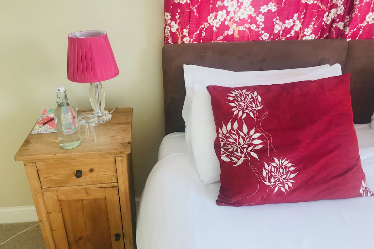 Un letto rifatto con cuscini bianchi e rossi, un comodino in legno con una lampada rosa e una bottiglia d'acqua.
