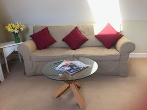 A beige sofa with two red pillows, a glass coffee table with magazines, and a vase of yellow flowers on a white side table. - Rosebank Bed & Breakfast (Etchingham)