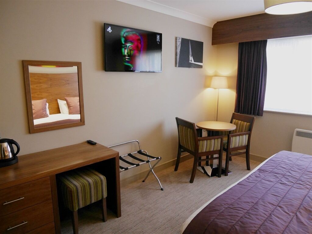 A hotel room with a bed, a desk with a chair, a small table with a chair, a TV mounted on the wall, and a lamp.