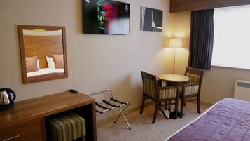 A hotel room with a bed, a desk with a chair, a small table with a chair, a TV mounted on the wall, and a lamp.