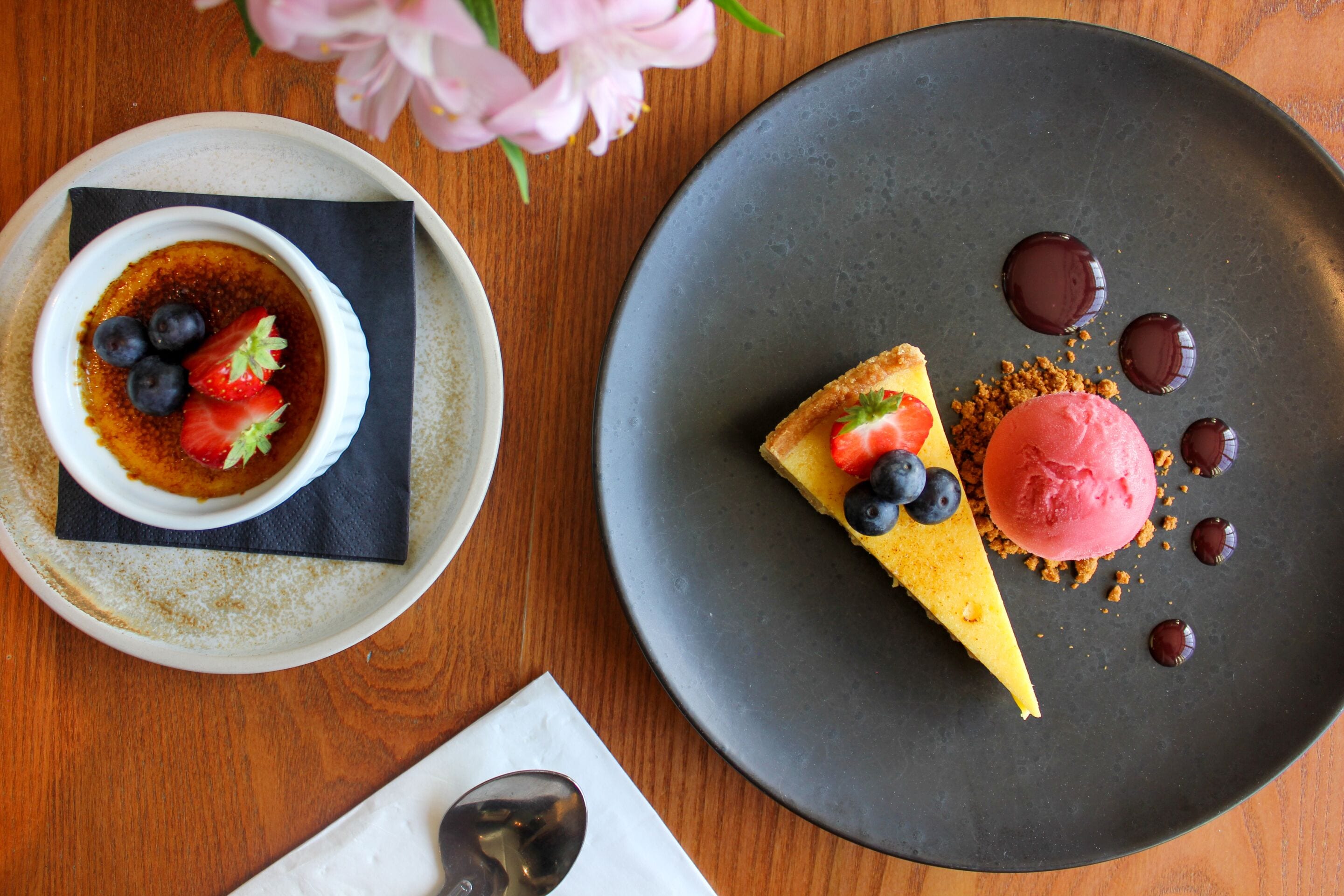 A dessert plate with a slice of cheesecake and ice cream, a bowl of crème brûlée with strawberries and blueberries, and a spoon on a napkin.