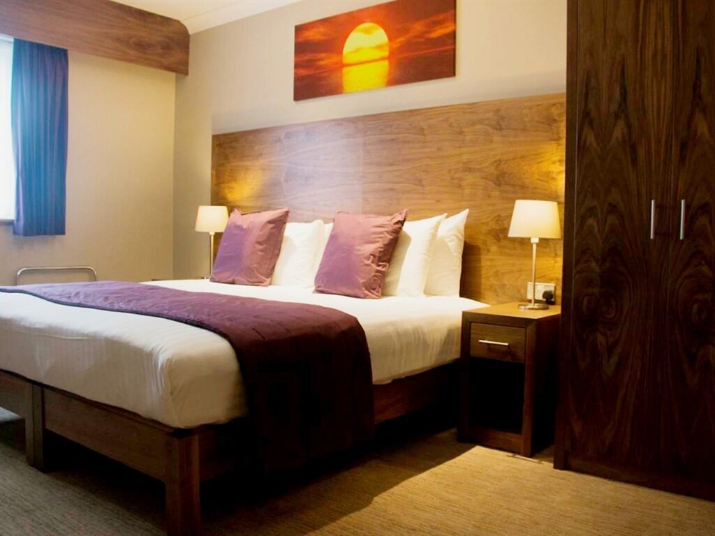 Een hotelkamer met een groot bed, twee nachtkastjes, een houten hoofdeinde en een schilderij van een zonsondergang.
