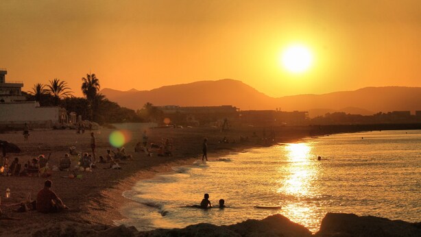 Escena de playa con gente, palmeras y montañas al fondo durante el atardecer.