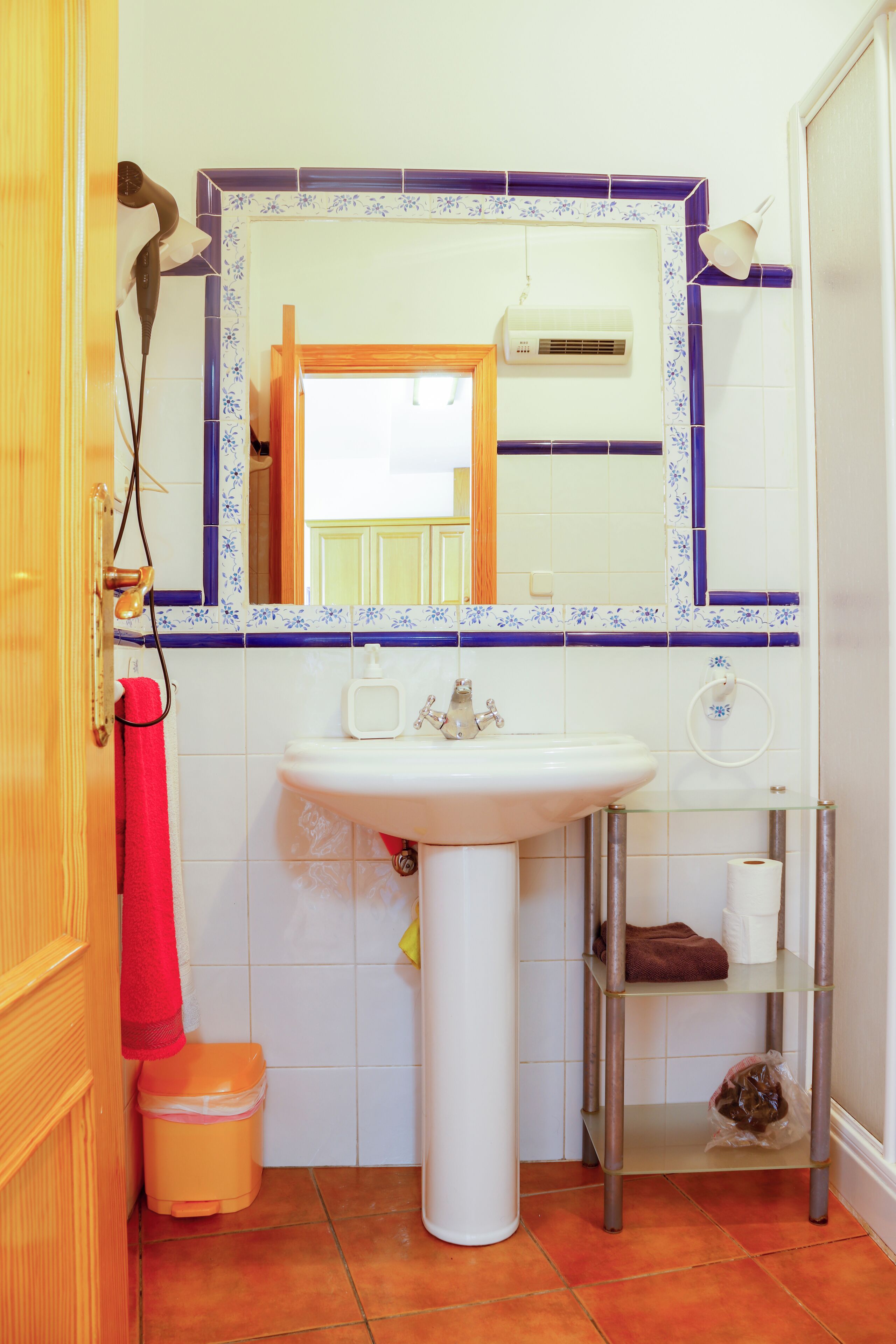 Een badkamer met een witte wastafel, een spiegel met blauwe lijst, een handdoekhouder, een prullenbak en een plank met toiletartikelen.
