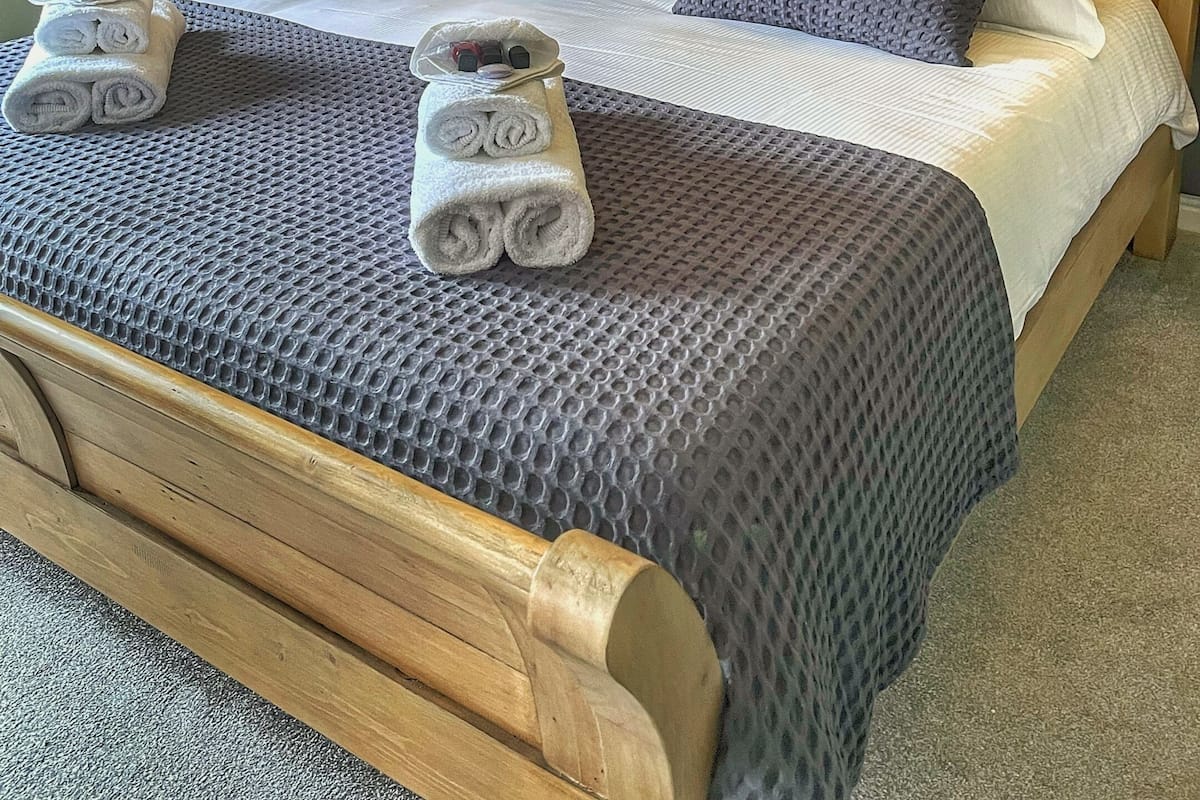 Uma cama bem arrumada com colcha cinza texturizada e almofadas, uma cabeceira de madeira e uma pequena mesa de cabeceira com uma luminária.