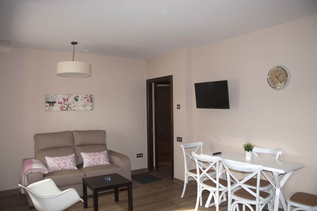 Uma sala de estar com sofá, mesa de centro, mesa de jantar com cadeiras, televisão fixada na parede e uma porta que dá para outro cômodo.