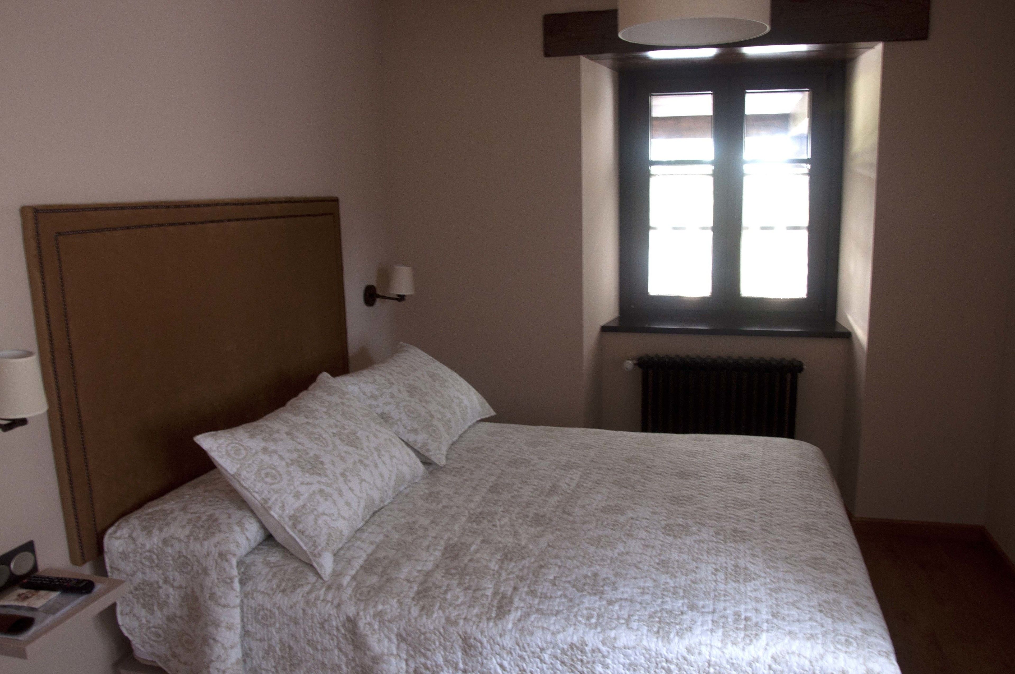 Une chambre à coucher comprenant un lit, une table de chevet, une lampe fixée au mur et une fenêtre avec des stores.