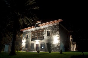 Edificio de piedra con un amplio ventanal y un balcón, rodeado de árboles y un césped bien cuidado.