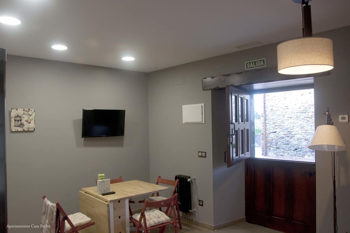 Uma área de refeições com mesa e cadeiras de madeira, televisão montada na parede e placa indicando a saída.