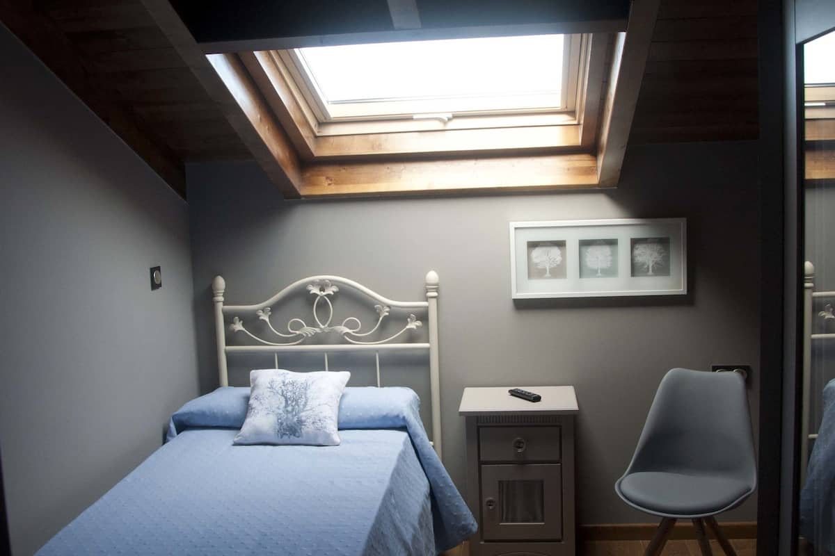 Quarto com claraboia, cama com cabeceira, mesa de cabeceira, uma cadeira e um pequeno armário.