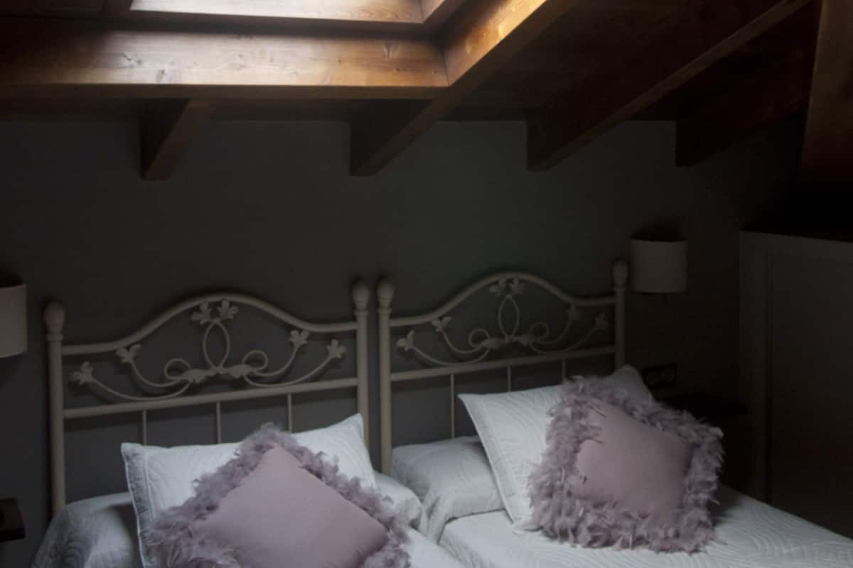 Quarto com claraboia, cama com roupa de cama branca e duas almofadas, e cabeceira branca.