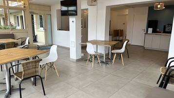 A modern dining area with wooden tables and chairs, a kitchen in the background, and a large window providing natural light.
