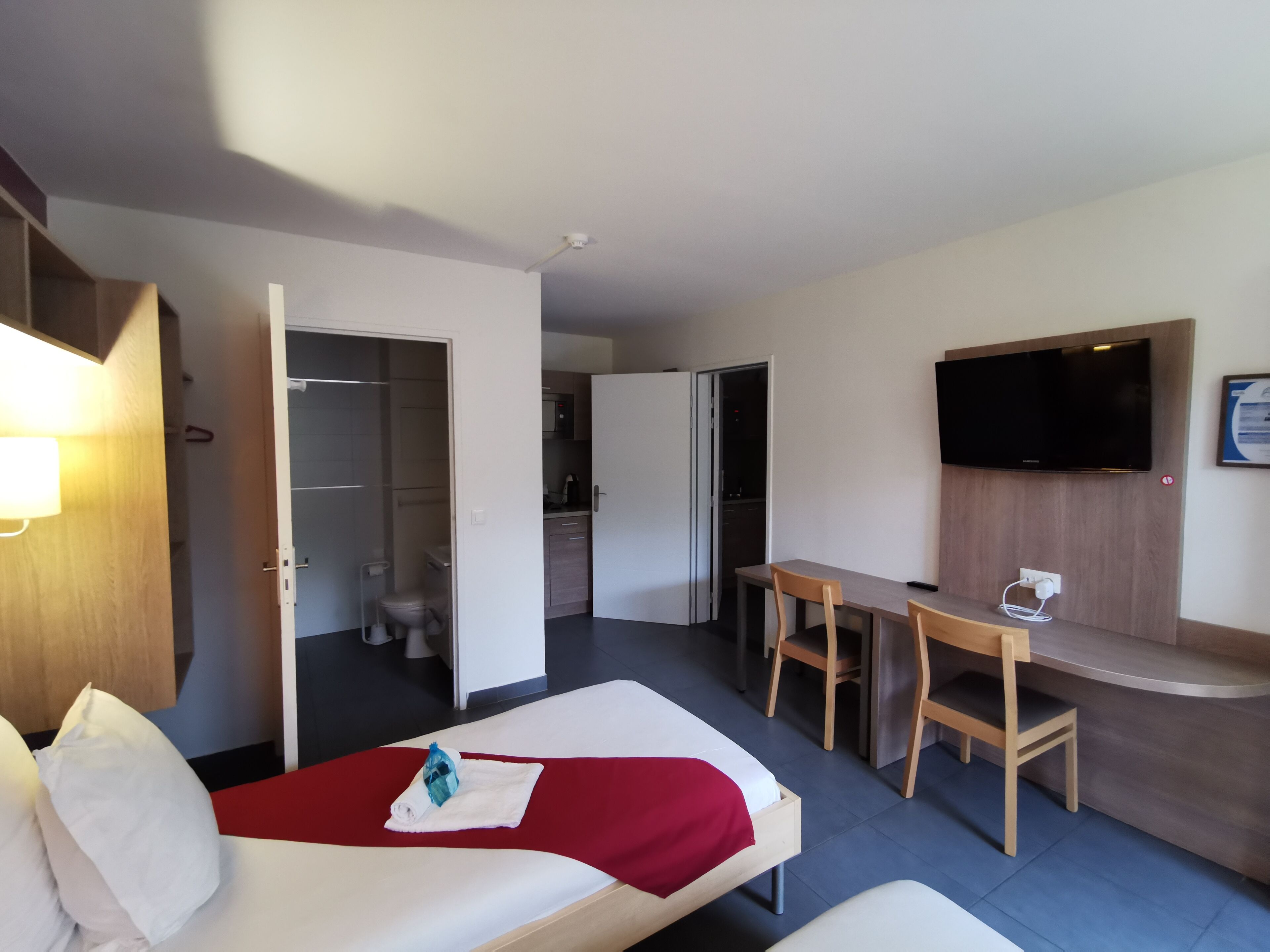 Quarto de hotel com cama, escrivaninha, televisão e armário.