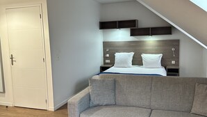 A modern bedroom with a lofted ceiling, a grey sofa, a wooden coffee table, and a bed with white linens.