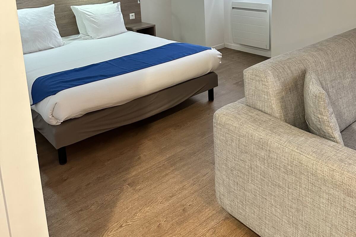 A bedroom with a bed, a nightstand, a built-in shelf, a window with curtains, and a sofa.