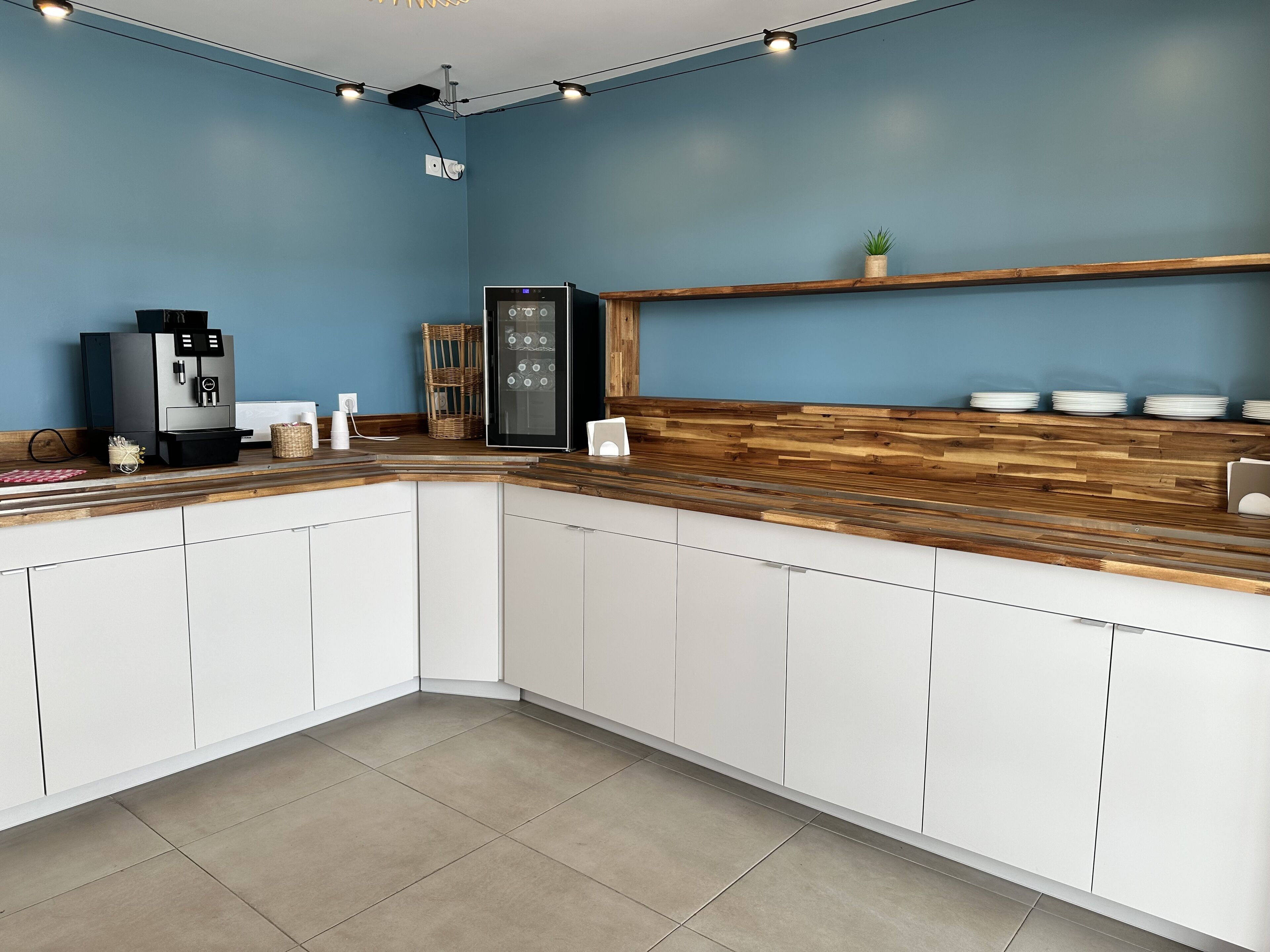 A modern kitchen with white cabinets, a wooden countertop, a coffee machine, a refrigerator, and a shelf with plates.
