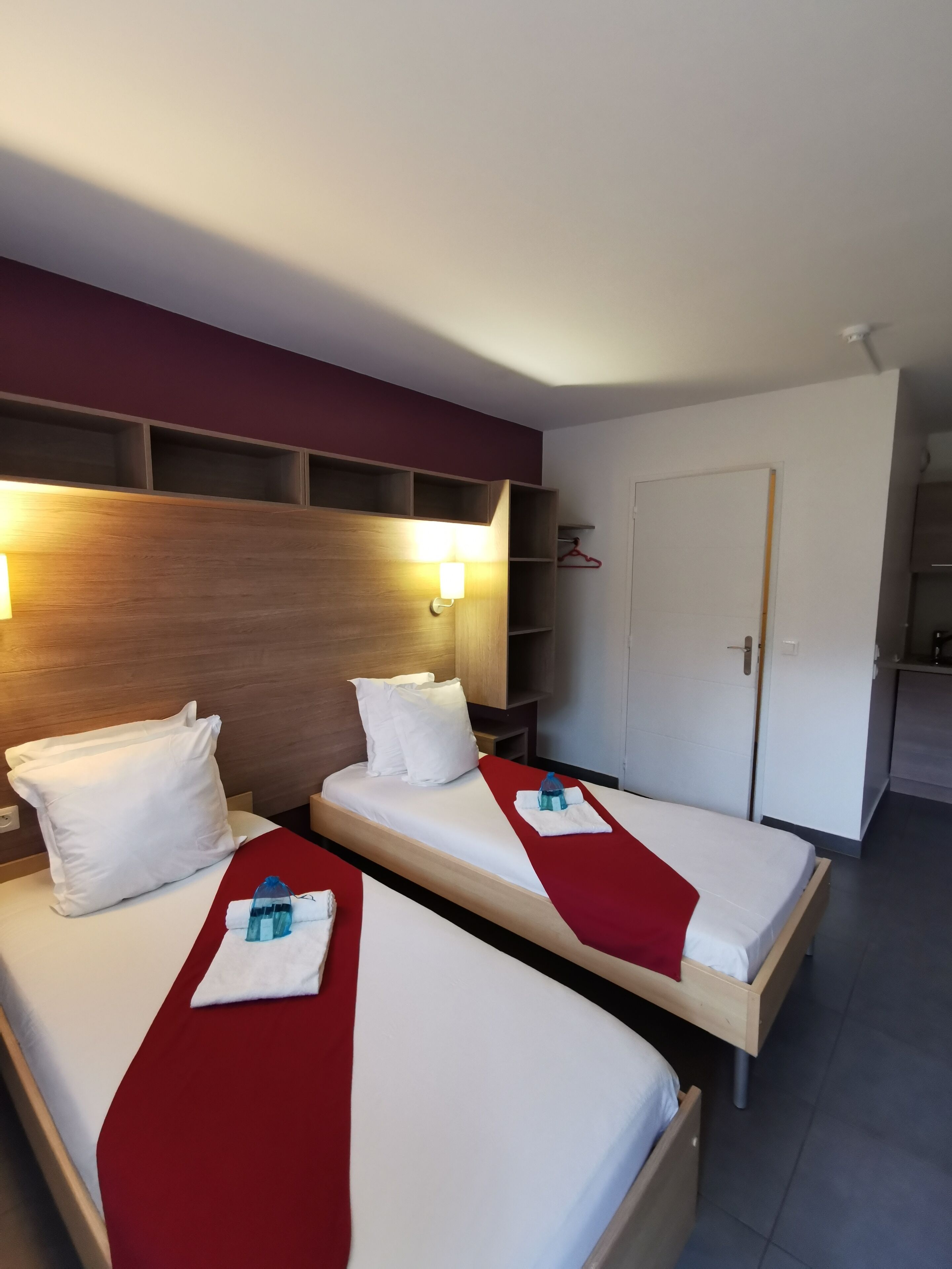 A hotel room with two beds, each with white linens and red bedspreads. There are wall-mounted light fixtures above the beds and a coat rack to the right.