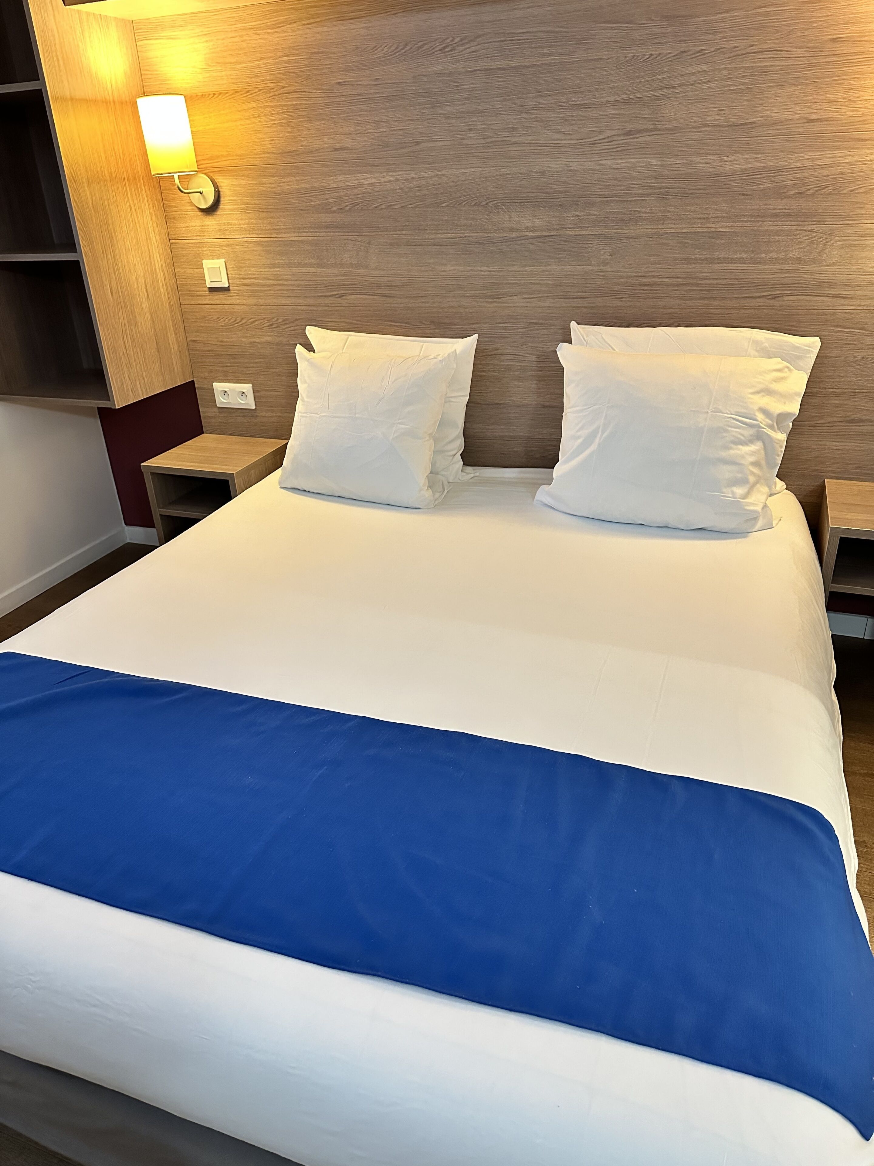 A neatly made bed with white bedding and a blue accent, a wooden headboard, and a wall-mounted light fixture.