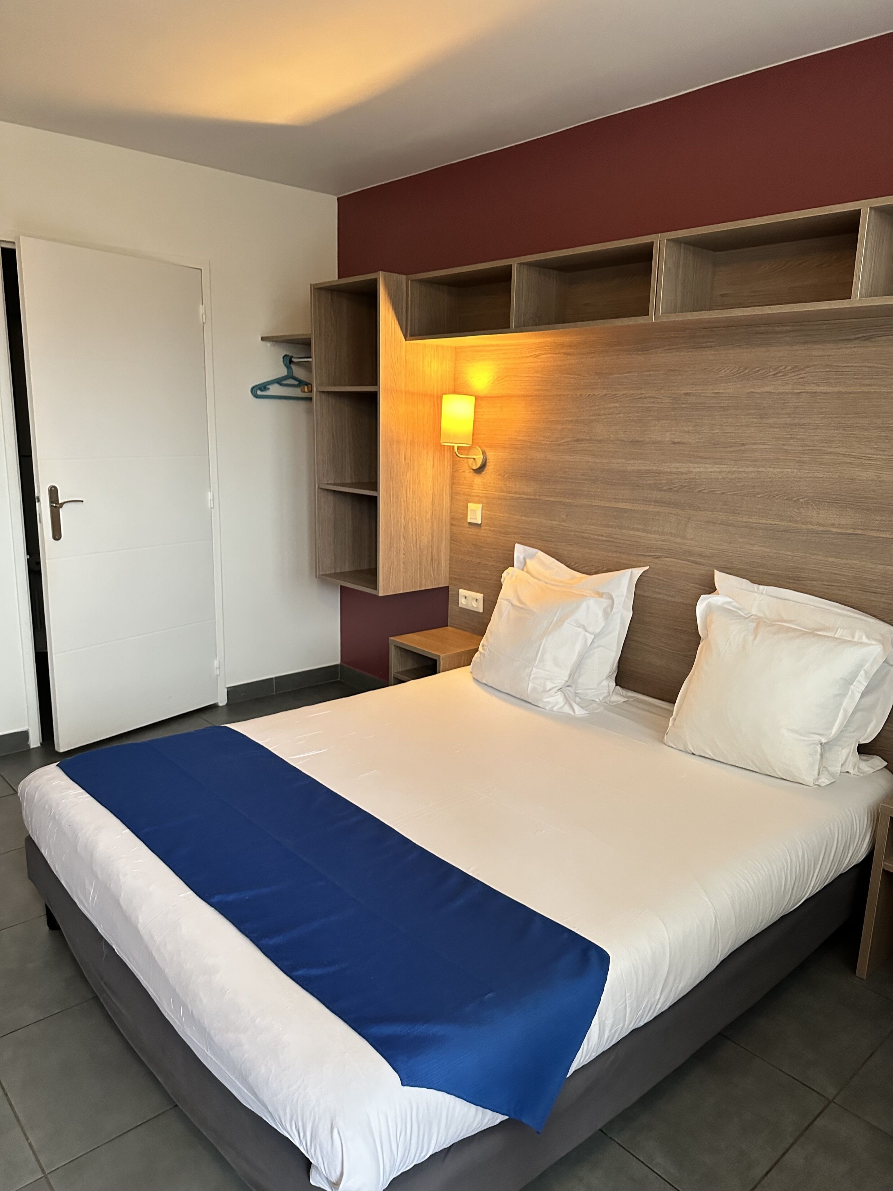 Ett hotellrum med en stor säng, en sänggavel i trä, en vägghylla och en sänglampa.