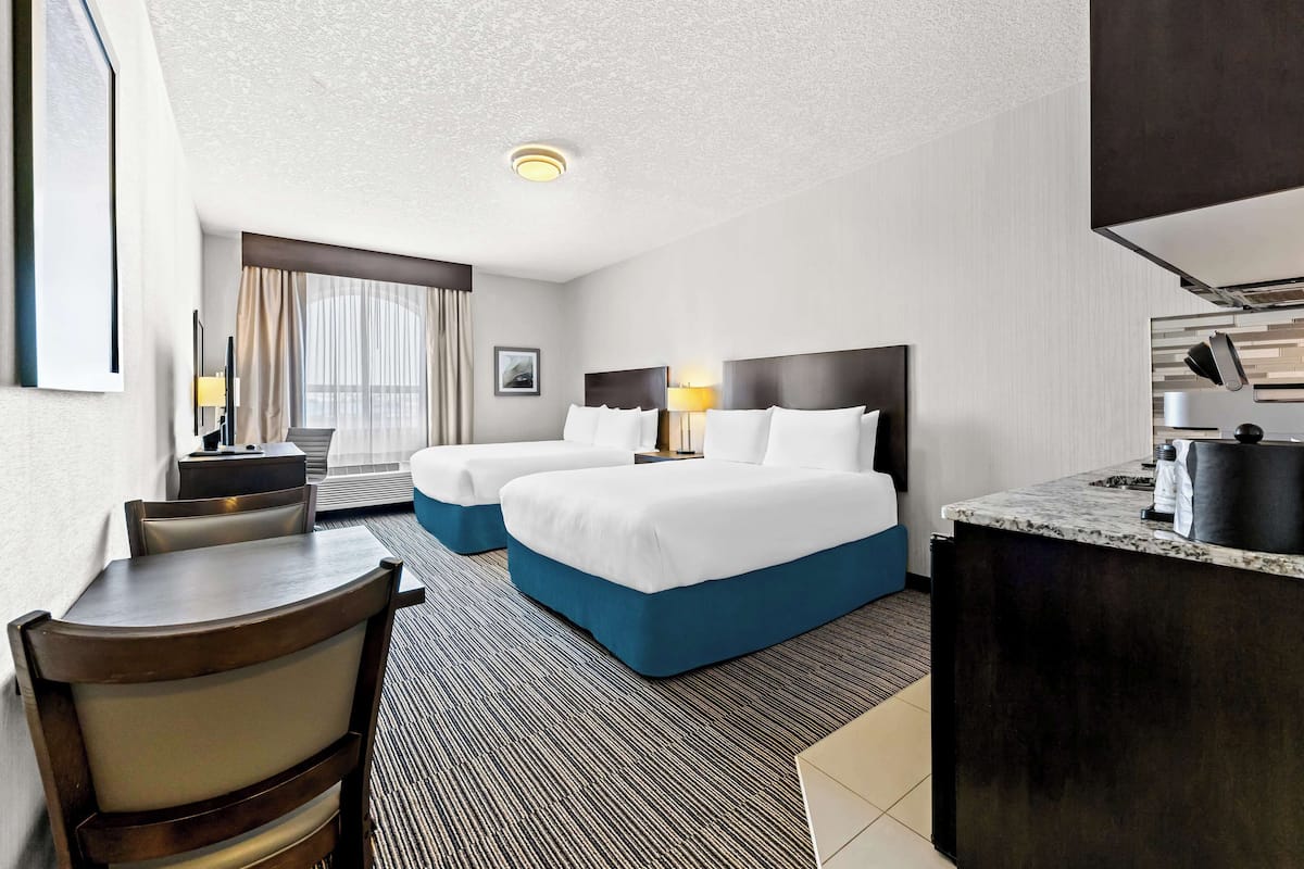 A hotel room with two beds, a desk, a chair, a small table, a lamp, and a kitchenette.