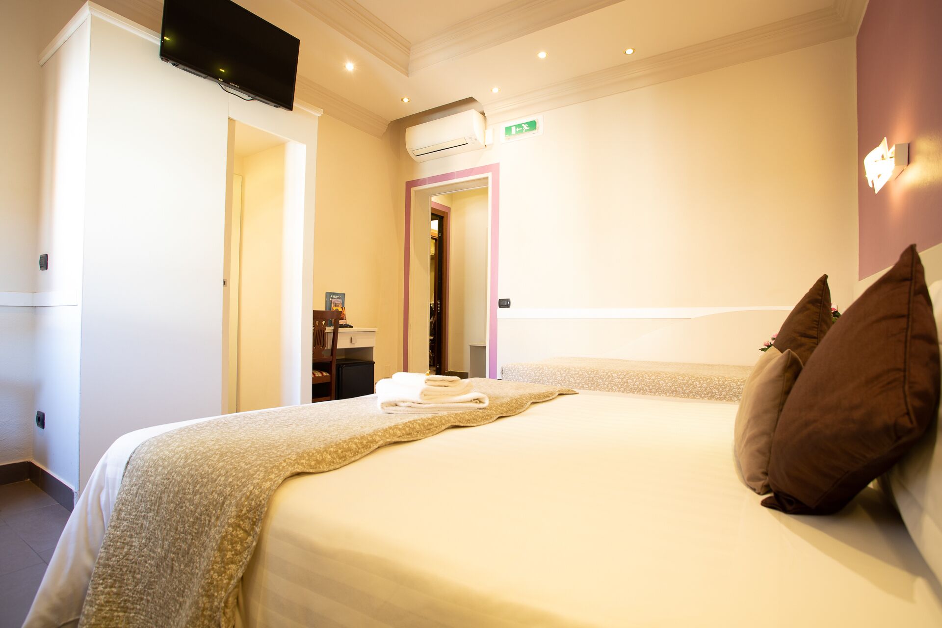 A hotel room with a bed, a flat-screen TV mounted on the wall, an air conditioning unit, and a door leading to a bathroom.