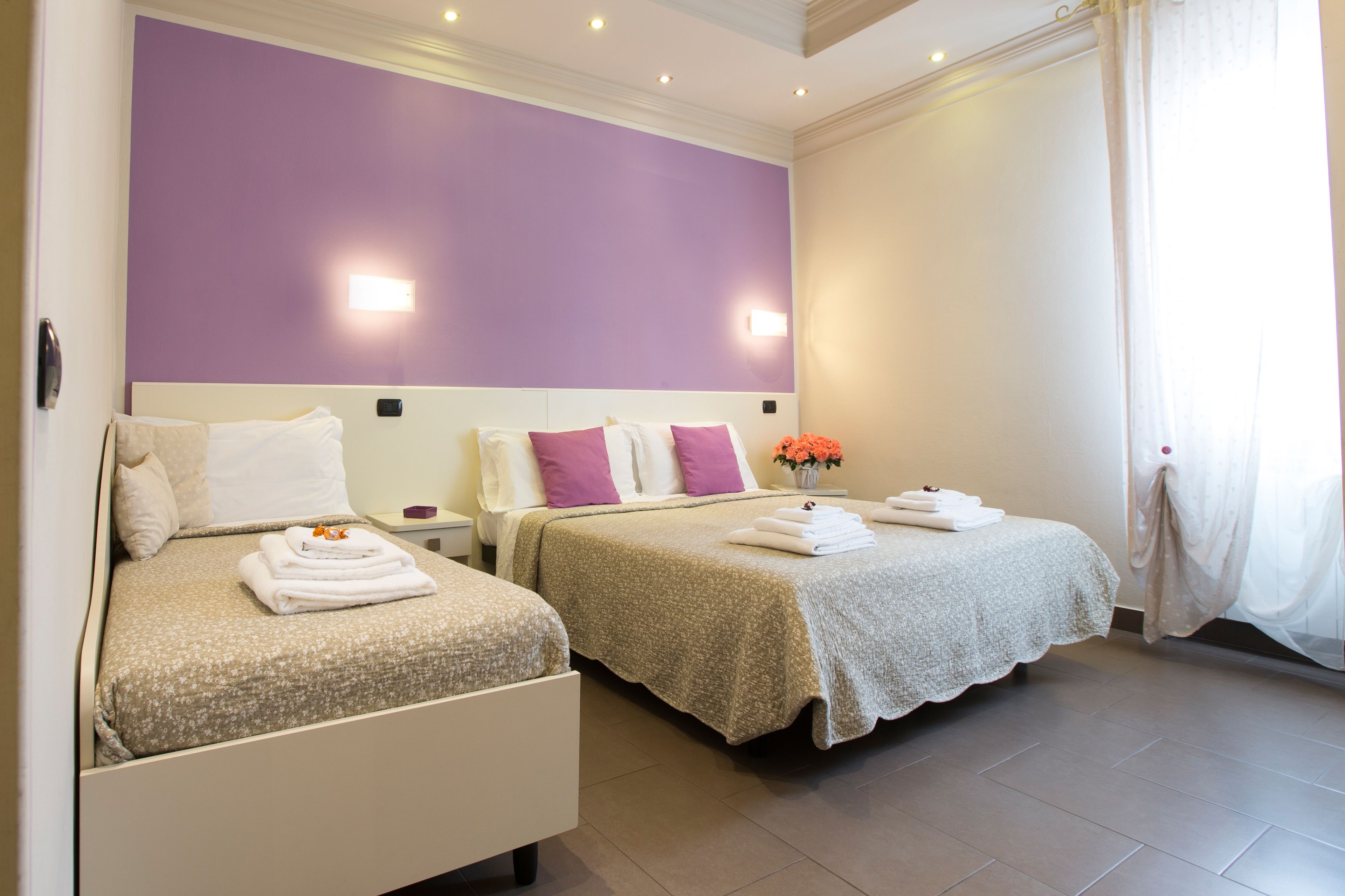 Ein Hotelzimmer mit zwei Betten, jeweils mit weißem Kopfteil und einem blumigen Überwurf. Es gibt zwei Nachttische, mit Lampen und auf einem davon eine Vase mit Blumen. Der Raum hat eine violette Akzentwand und ein großes Fenster mit weißen Vorhängen.