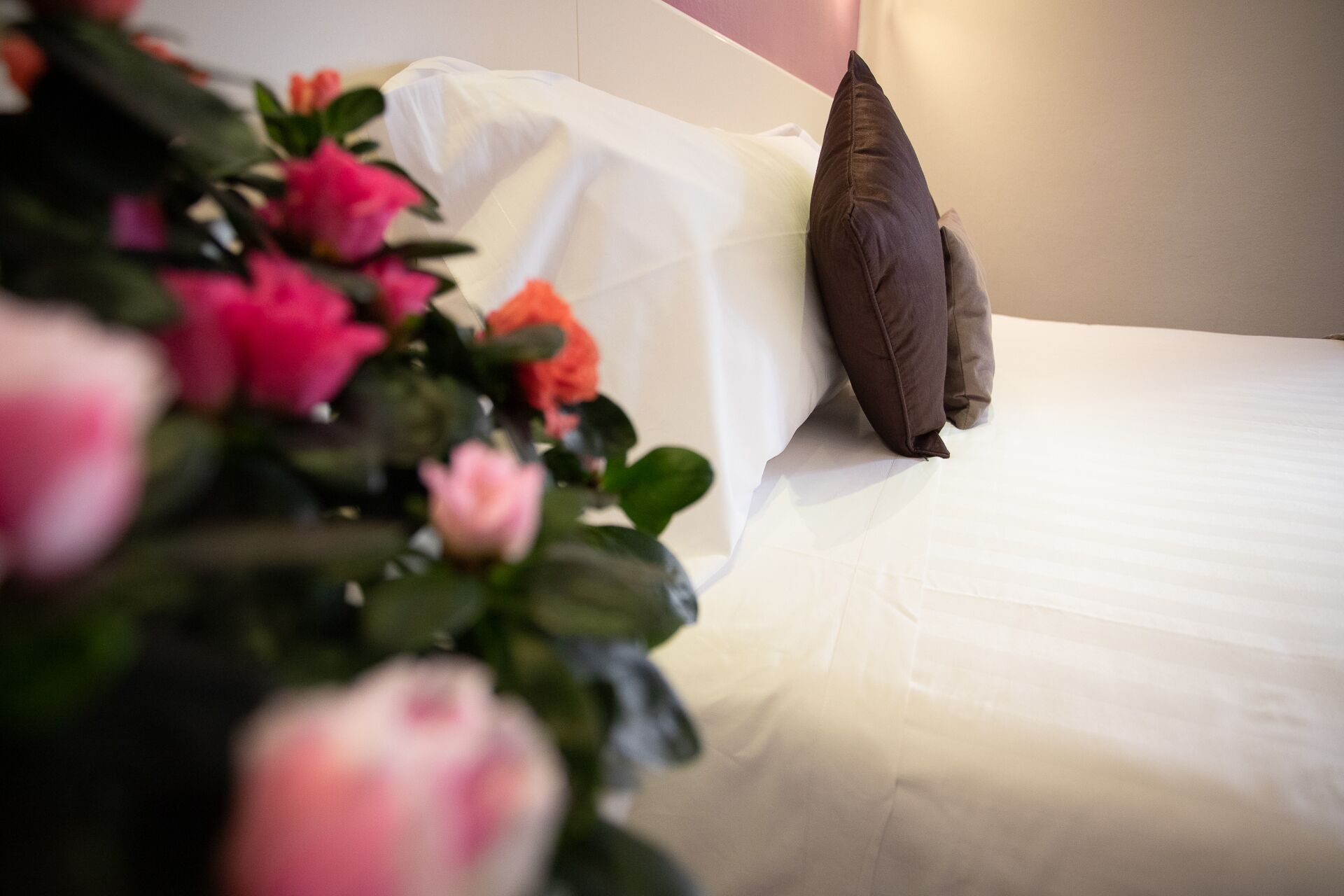 Una cama con sábanas blancas y almohada marrón, rodeada de flores rosas y anaranjadas.