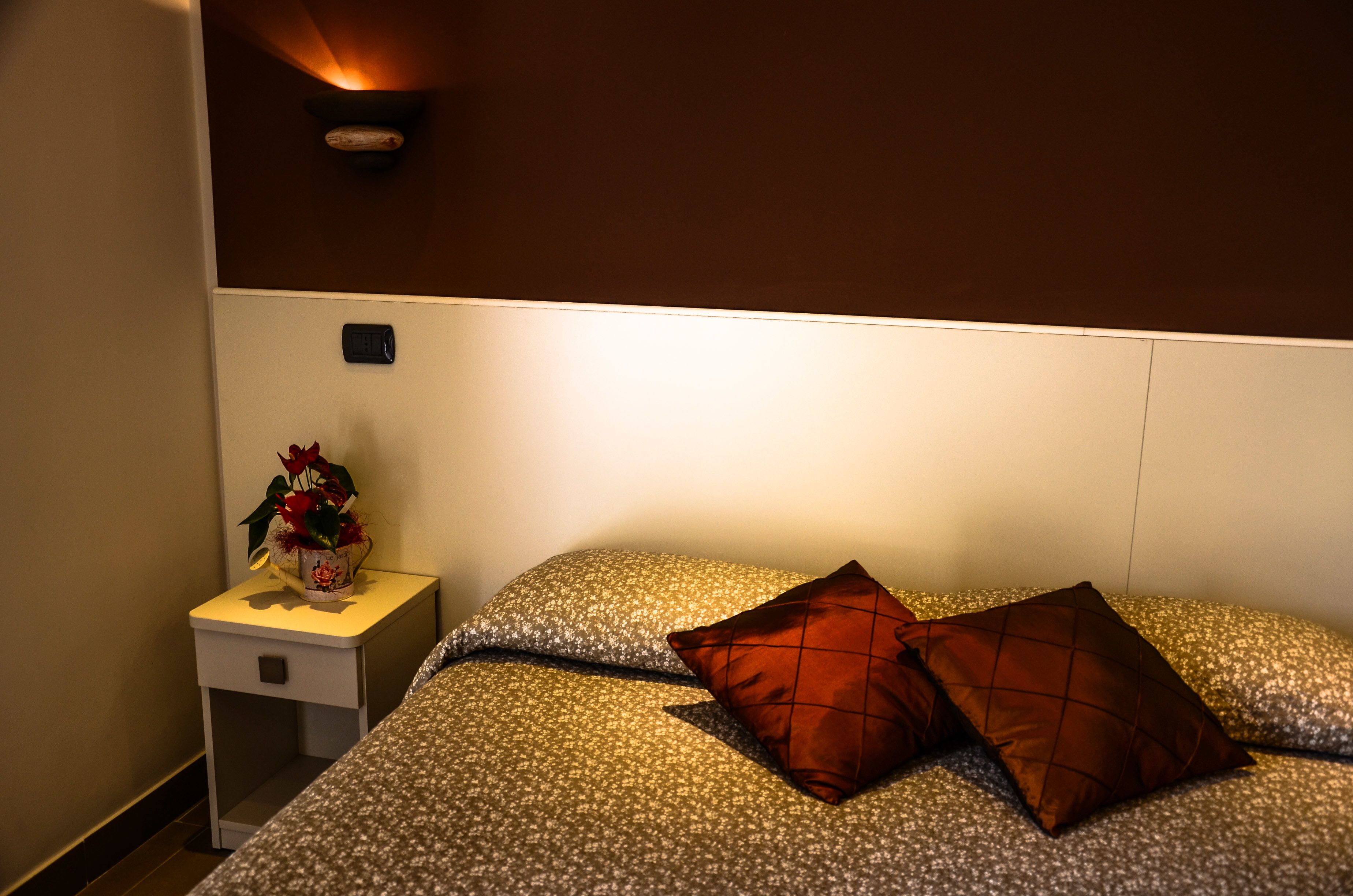 Een net opgemaakt bed met behangpatroon, een klein nachtkastje met een bloemenvaas, en een wandlamp.