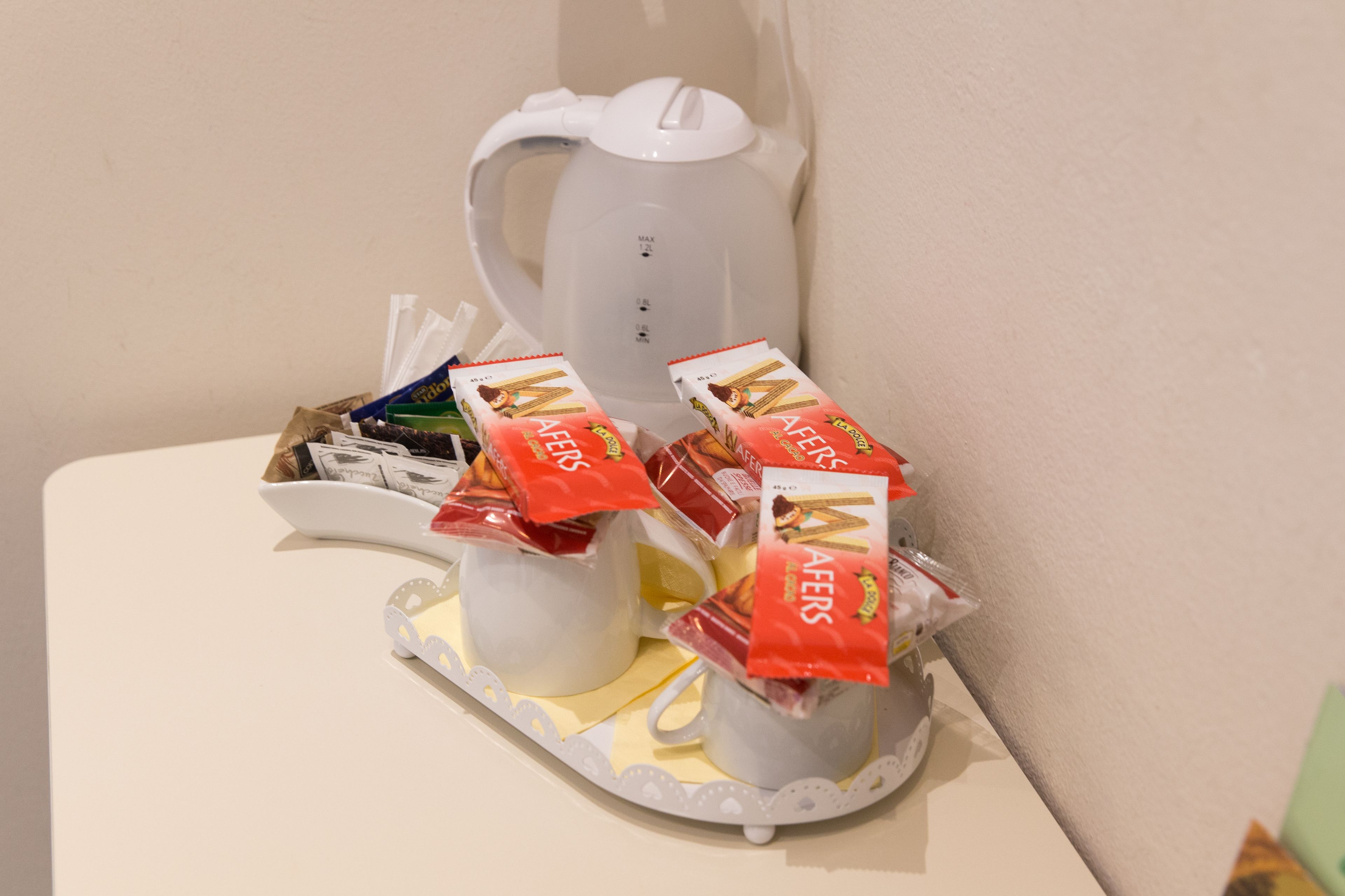 Valkoinen pöytä, jonka päällä on valkoinen teekannu ja kaksi valkoista kuppia. Jokaisen kupin päällä on punainen paketti, jossa lukee teksti ”WAFFERS”.