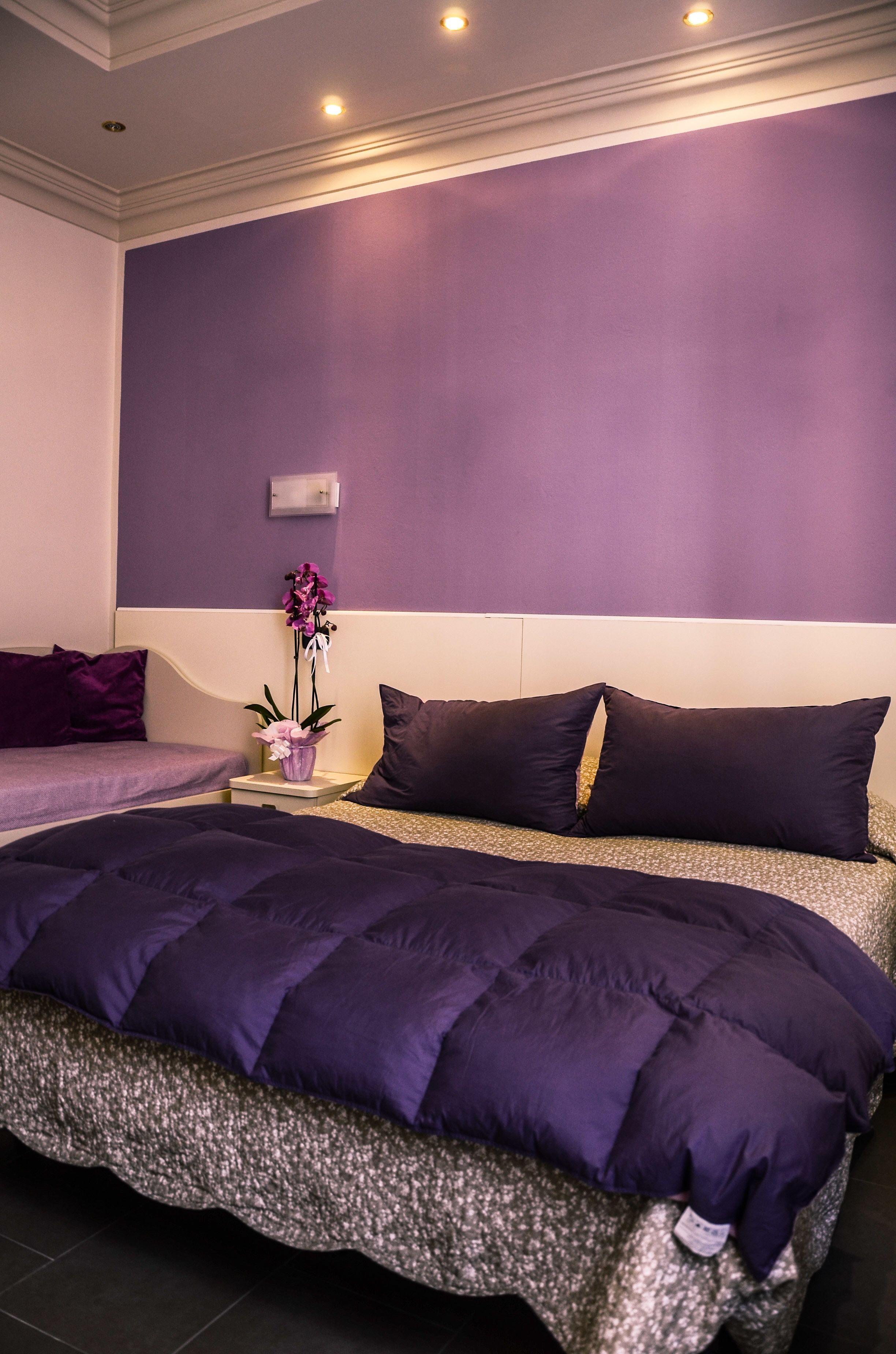 A bedroom with a large bed, purple bedding, a nightstand with a vase of flowers, and a wall-mounted light fixture.