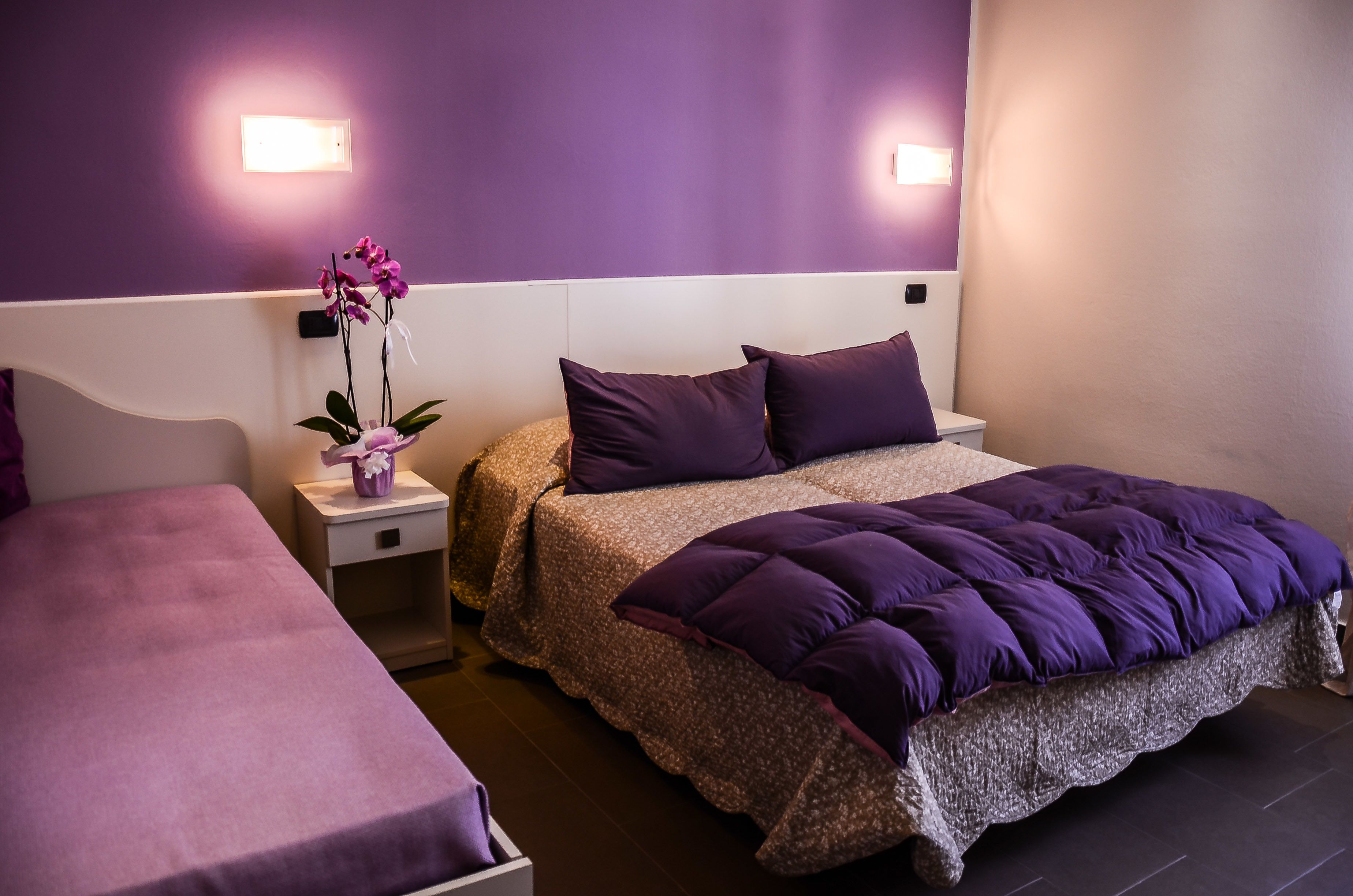 A hotel room with two beds, purple bedding, a nightstand with a vase of flowers, and wall-mounted lights.