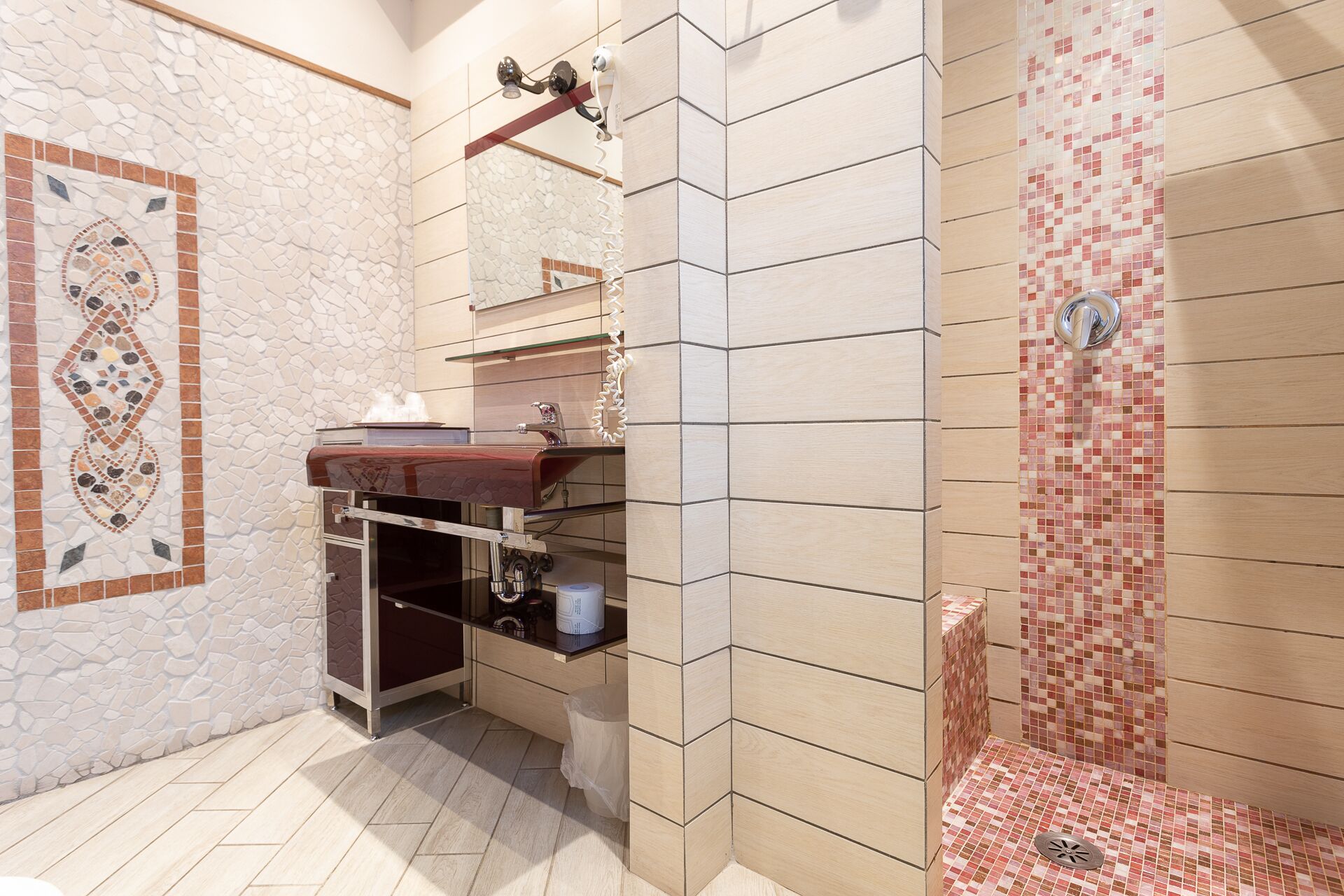 A bathroom with a stone mosaic wall, a wooden vanity, and a tiled shower area.