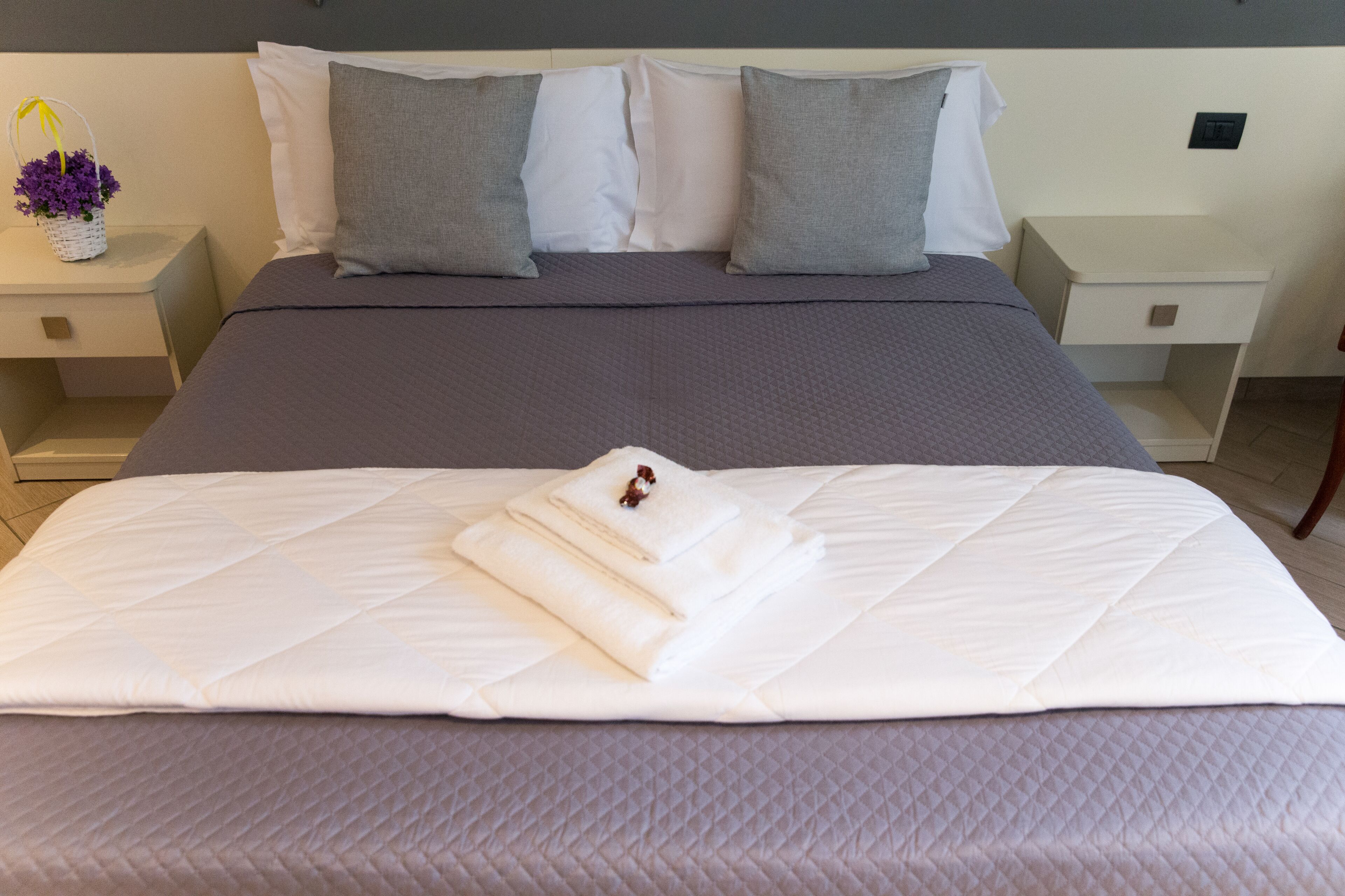 A neatly made bed with a white comforter and pillows, a small bedside table with a basket of flowers, and a nightstand with a lamp.