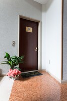 Entrada de hotel con puerta de madera, planta en maceta con flores rosas y felpudo.
