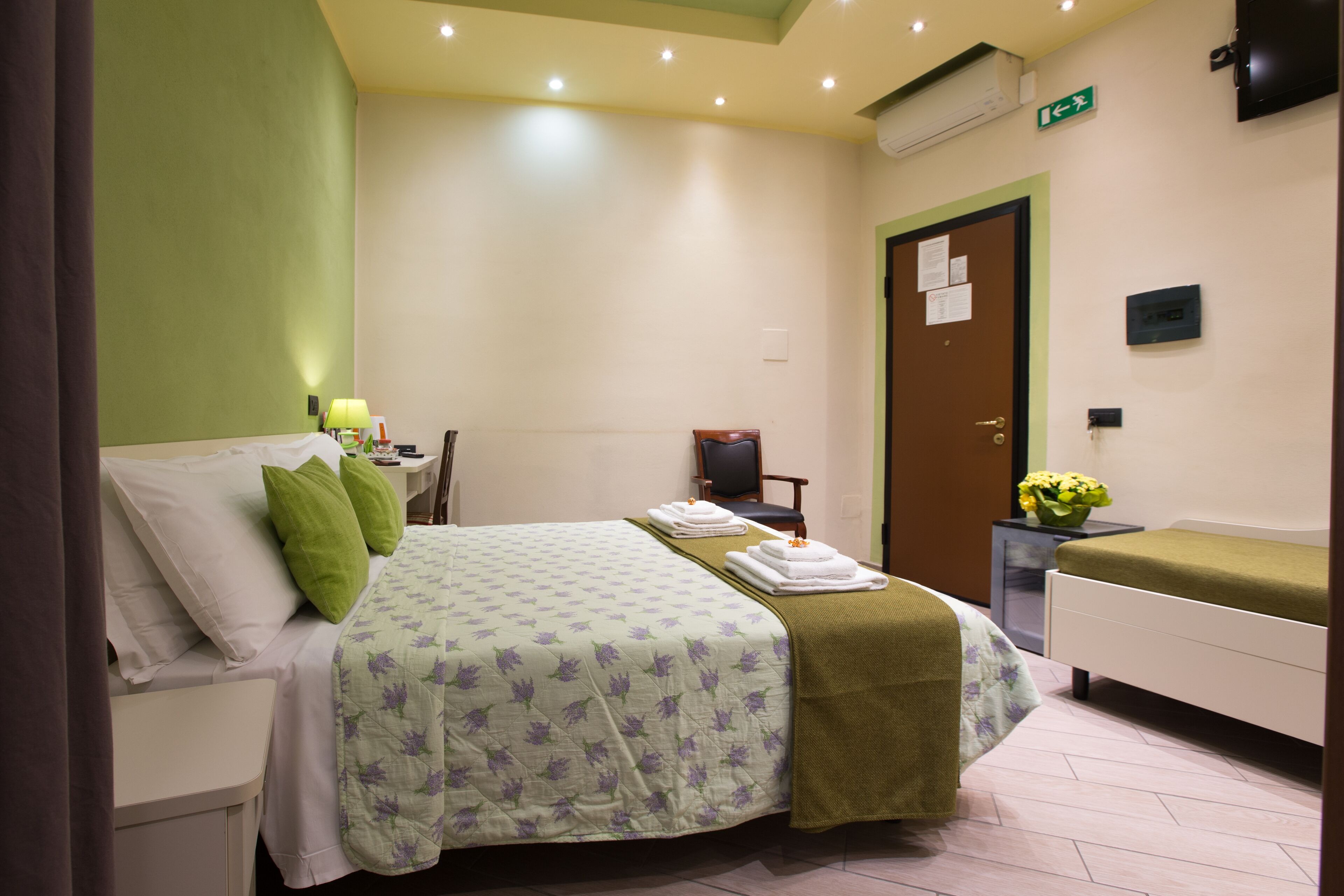 A hotel room with a bed, a chair, a small table with a vase of flowers, and a bedside table with a lamp.